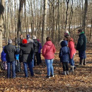 A group of adults and youth in spring outdoor clothes gathered around a guide in front of a stand of birch trees.