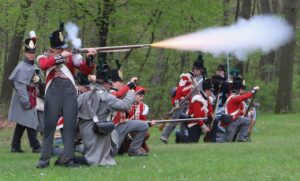 Men in red coats and shako hats participating in a reenactment of a battle from the War of 1812.